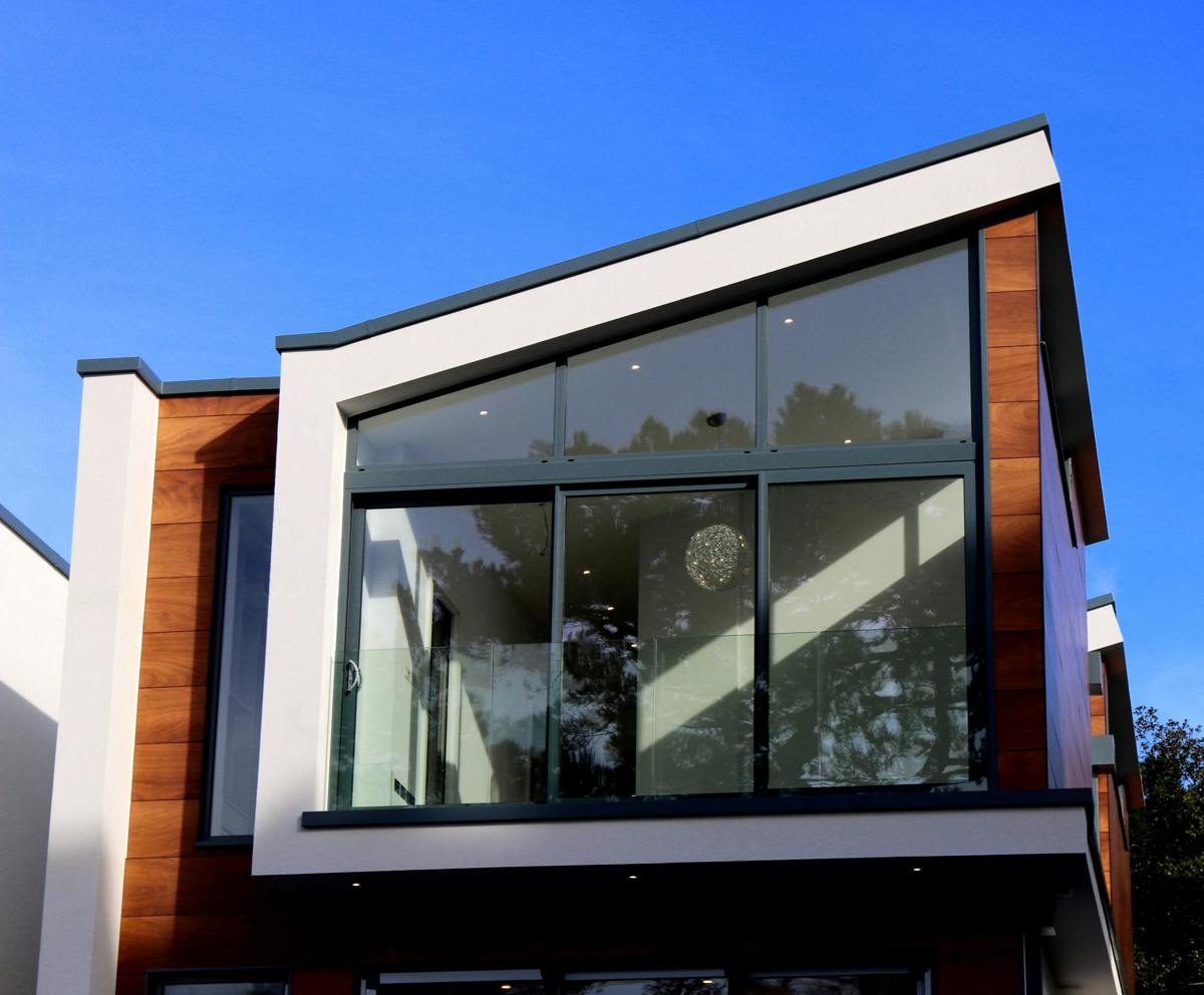 Modern tvåplans villa med stora glaspartier, träpanel och vit putsfasad mot blå himmel.