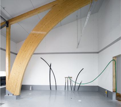 Träbalk i bågformad konstruktion i en byggnad med vitmålade väggar och grått golv.