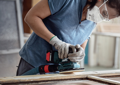 Person slipar trä med en elektrisk slipmaskin, iförd blå tröja, handskar och ansiktsmask.