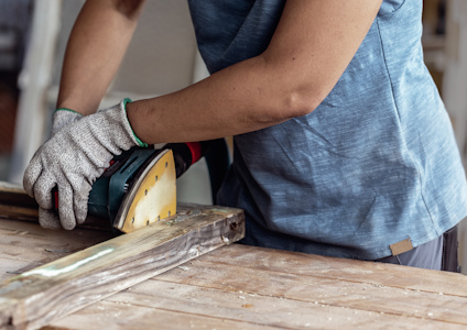 Person som slipar ett träbräde med en elektrisk slipmaskin, iförd arbetshandskar och blå tröja.