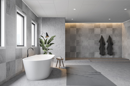 Modernt badrum med fristående vit badkar, grå väggplattor och inomhusväxt bredvid. Flera fönster och två mörka handdukar.