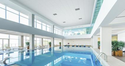 Inomhuspool med blått vatten, omgiven av vita väggar och solstolar, stora fönster runt omkring.