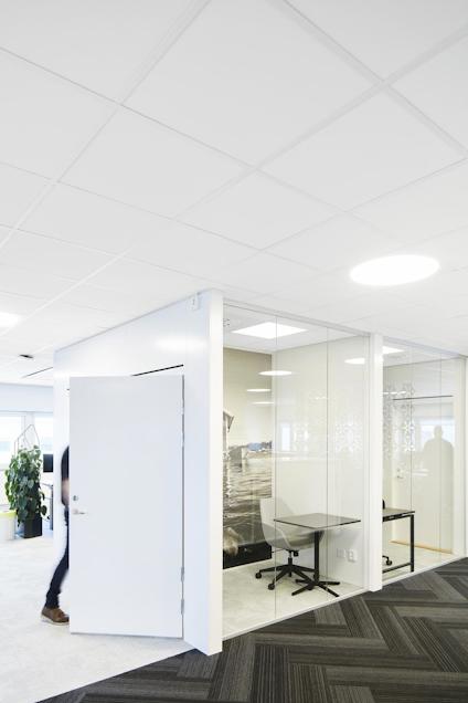 Kontor med glasväggar, öppen dörr, skrivbord och stolar. Ljust tak med takplattor och infällda lampor. Golv med mörk matta.