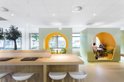 Modernt kontorsutrymme med trämöbler, vita stolar, inomhusträd och runda, gula nischer med människor.
