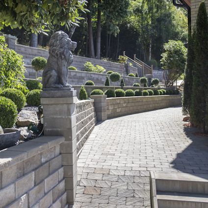 Slingrande stenlagd gångväg med skulptur av en lejonstaty. Omgiven av stenmur och prydligt klippta buskar.