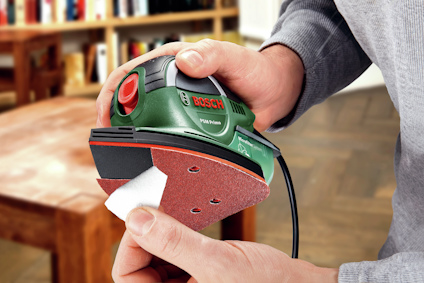 Person håller i en grön och röd Bosch-slipmaskin, modell PSM Primo, med en trekantig slipyta.