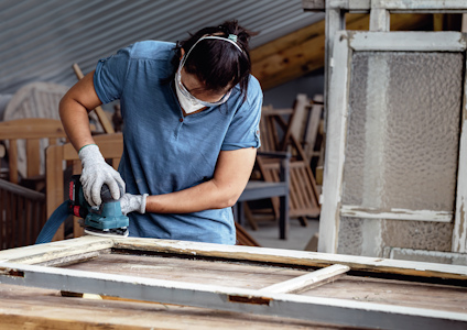 En person slipar en träram med en elektrisk slipmaskin, iförd skyddsmask och handskar.