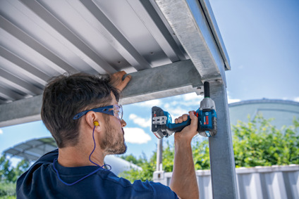 Person skruvar upp en metallpanel med trådlös borrmaskin, bär skyddsglasögon och hörselskydd.