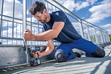 En person i arbete klädd i skyddsglasögon och hörselproppar, använder en batteridriven borrmaskin på ett metallstängsel.