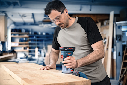 En person slipar en träbänk med en elektrisk slipmaskin i en verkstad, omgiven av hyllor och verktyg.