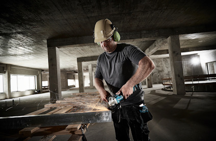 Person i skyddshjälm och hörselskydd slipar metallbalk i en betongbyggnad.