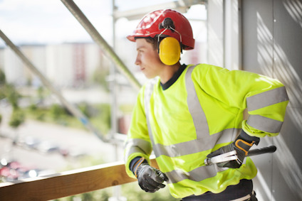 Person i reflexjacka och hjälm blickar ut från en byggnadsställning, med hörselskydd och handskar.