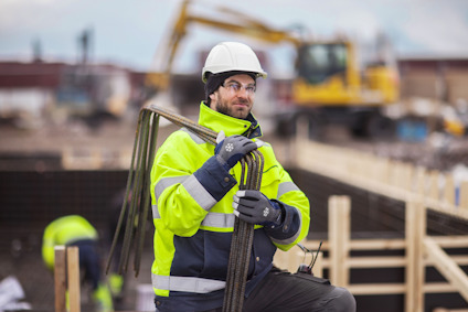 Person i reflexjacka och hjälm håller i armeringsjärn på en byggarbetsplats med grävmaskin i bakgrunden.