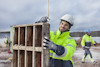 Person i hjälm och reflekterande jacka använder en måttstock för att mäta träform medan andra arbetar i bakgrunden.