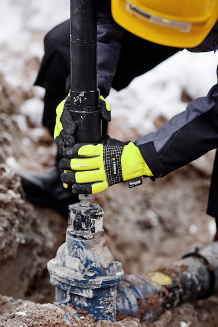 Person med gula handskar och hjälm arbetar med ett rör vid en byggarbetsplats.
