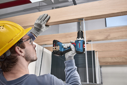 Person i skyddshjälm och glasögon använder en blå, sladdlös borrmaskin på träbalkar inomhus.