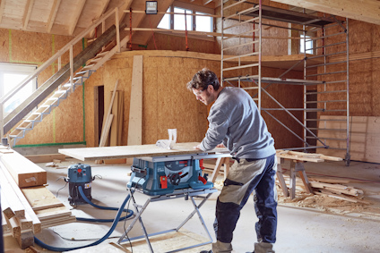 En person arbetar vid en elektrisk såg i ett halvt färdigt byggprojekt med träbjälkar, stege och ställningar i bakgrunden.