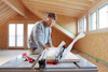 Man using table saw to cut wood planks in an attic under construction. Wearing safety glasses and casual clothing.