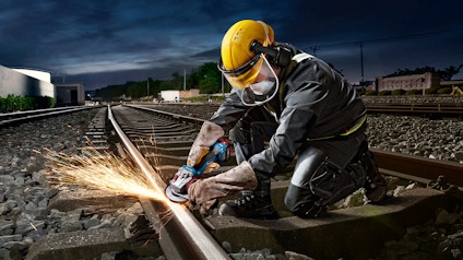 Person använder slipmaskin på järnvägsräls, bär skyddsutrustning inklusive hjälm och glasögon.