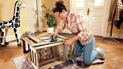 Person slipar en träpall på knä i ett rum med växt, dörr och giraff-figur.