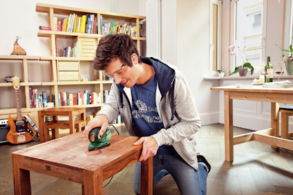 Man slipar bord med slipmaskin i ett vardagsrum. Bakom syns en bokhylla, ett bord och en gitarr.