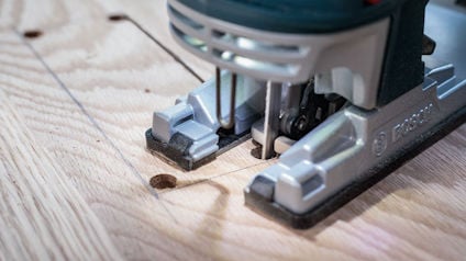 Elektrisk sticksåg som skär igenom ett trästycke med borrade hörnmarkeringar.