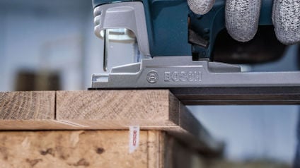 Sticksåg används för att såga trä. Märkt "BOSCH". En del av handen och handske synliga.