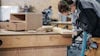 Person arbetande vid en arbetsbänk med träbearbetning, bär skyddsutrustning, verktyg och trästycken synliga.