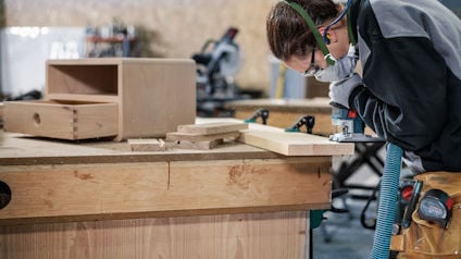 Person arbetande vid en arbetsbänk med träbearbetning, bär skyddsutrustning, verktyg och trästycken synliga.