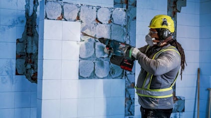 En person i skyddsutrustning tar bort kakel från en vägg med en borrhammare.
