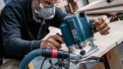 Person med ansiktsmask och skyddsglasögon använder en blå träfräs på träskiva.