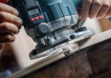 En person använder en handhållen träfräs på en träyta; mätningarna på digital displayen visar "390".