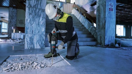 En person i säkerhetsutrustning använder en borrmaskin på ett betonggolv i en inomhusmiljö med marmorväggar och trappor.