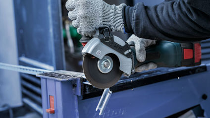 Person använder en vinkelslip för att kapa en metallstång. Handskar och skyddsjacka bärs.