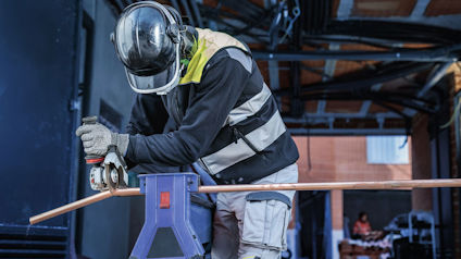 En person i skyddsutrustning bockar ett kopparrör med hjälp av en bockmaskin i en industriell miljö.