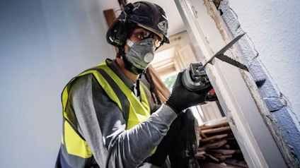 Person i skyddskläder och hjälm använder en eldriven såg för att skära i en dörrkarm.