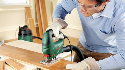 Person som använder en grön elektrisk sticksåg för att skära en träplanka på en arbetsbänk inomhus.