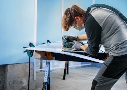 Person med skyddsglasögon och hörselskydd använder en elektrisk såg för att skära en träskiva på ett arbetsbord.