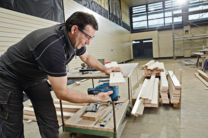 En person i skyddsglasögon slipar en träbräda med en elverktyg i en verkstad.