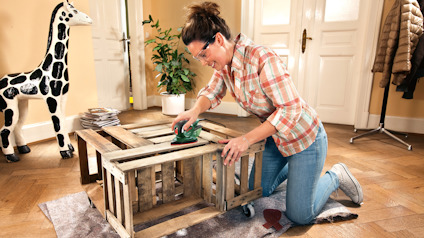 Kvinna slipar en träpall med verktyg inomhus, omgiven av leksaksgiraff och växter.