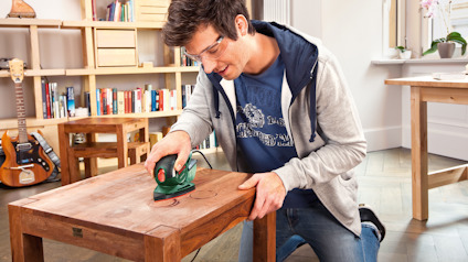 En person slipar en träbordsyta med en grön och svart slipmaskin i ett vardagsrum.