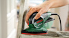 En hand håller i en grön och svart Bosch PSM Primo slipmaskin, används på en träram.