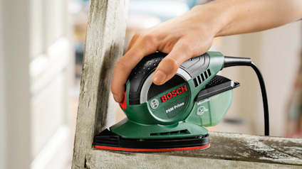 En hand håller i en grön och svart Bosch PSM Primo slipmaskin, används på en träram.