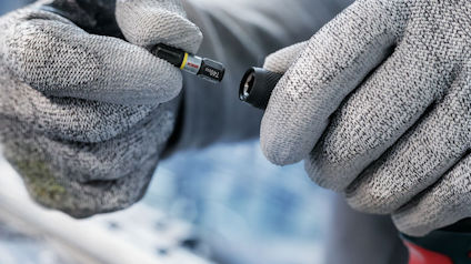 Händer i grå handskar håller ett litet verktyg eller komponent.