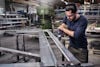 Person i skyddsglasögon borrar i metallprofil i verkstadsmiljö. Hyllor och maskiner i bakgrunden.