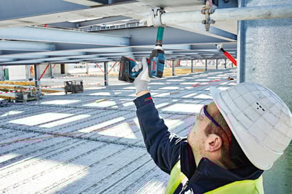 Byggarbetare med skyddshjälm och väst använder elektrisk borr på metallbalk.