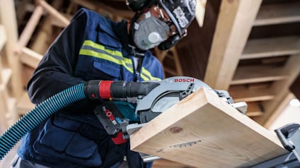 Person bär skyddsutrustning och använder en Bosch cirkelsåg för att såga trä i en byggmiljö.