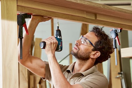 Person som använder en skruvdragare på träram, omgiven av klämmor, iklädd skyddsglasögon.