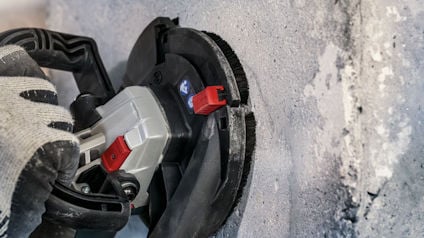 Hand som håller en vinkelslip mot betongvägg, med grå handske och röd detalj på verktyget.
