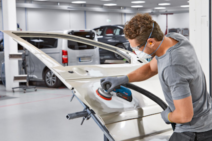 Person slipar en silverfärgad bildörr med en elektrisk slipmaskin i en verkstad.